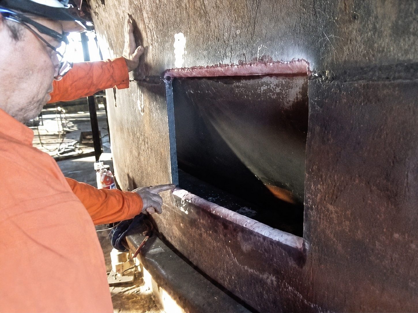 Worker in orange safety gear inspecting a rectangular opening in a large metal structure.