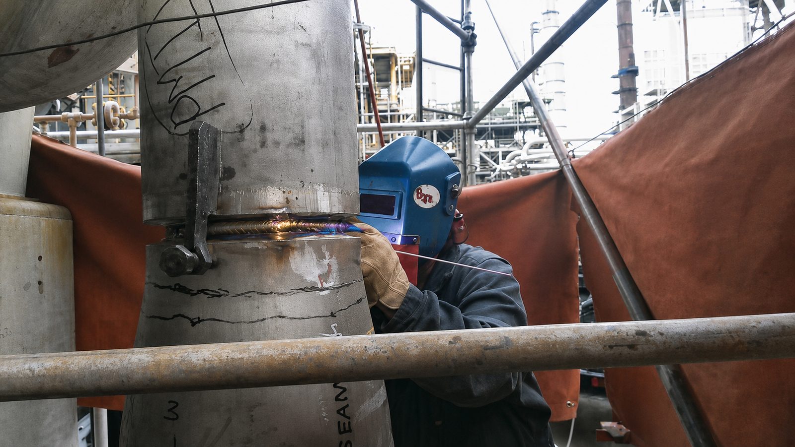 Welder working on a vertical pipe joint in an industrial setting.