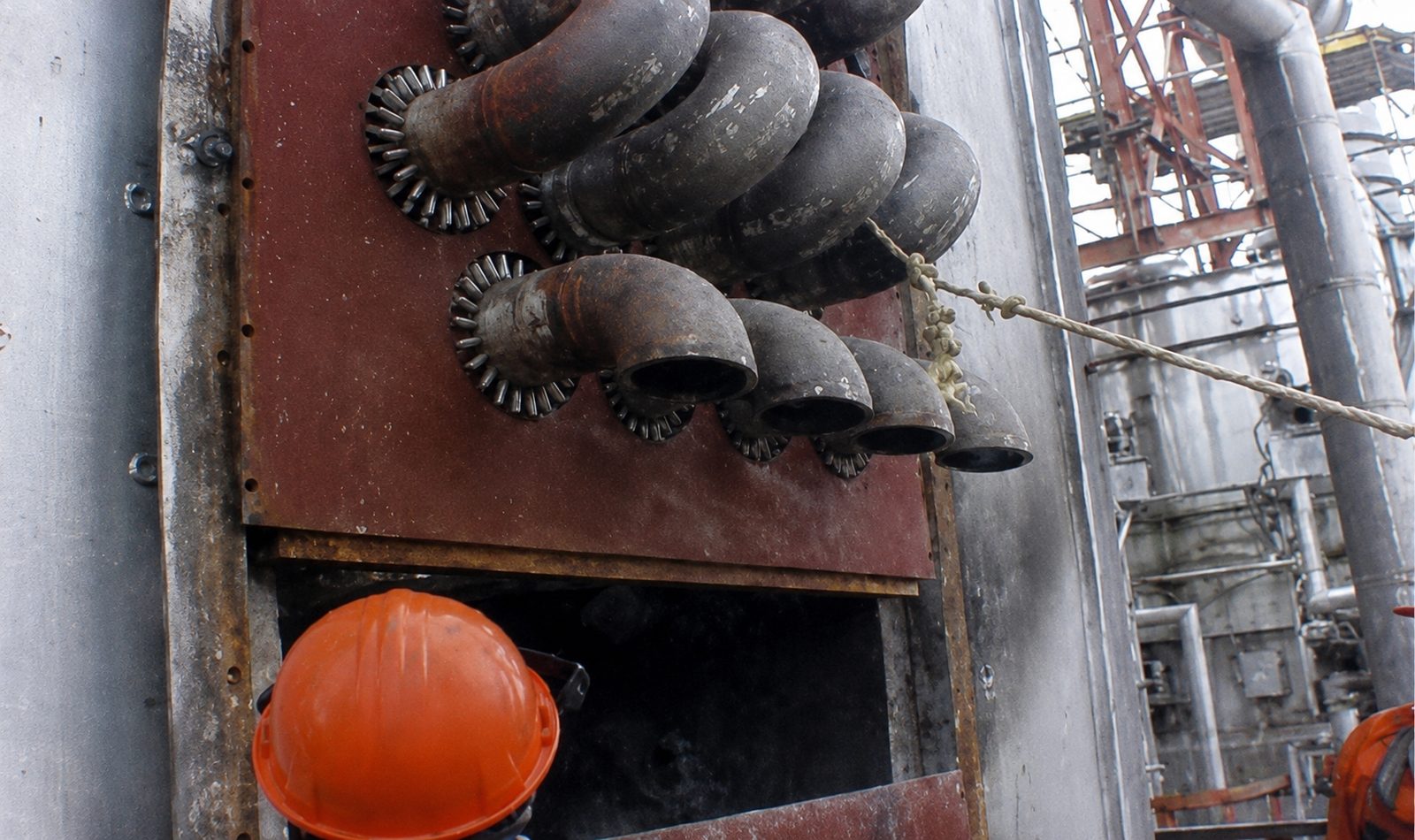 Industrial heater piping with a worker wearing an orange hard hat in the foreground.