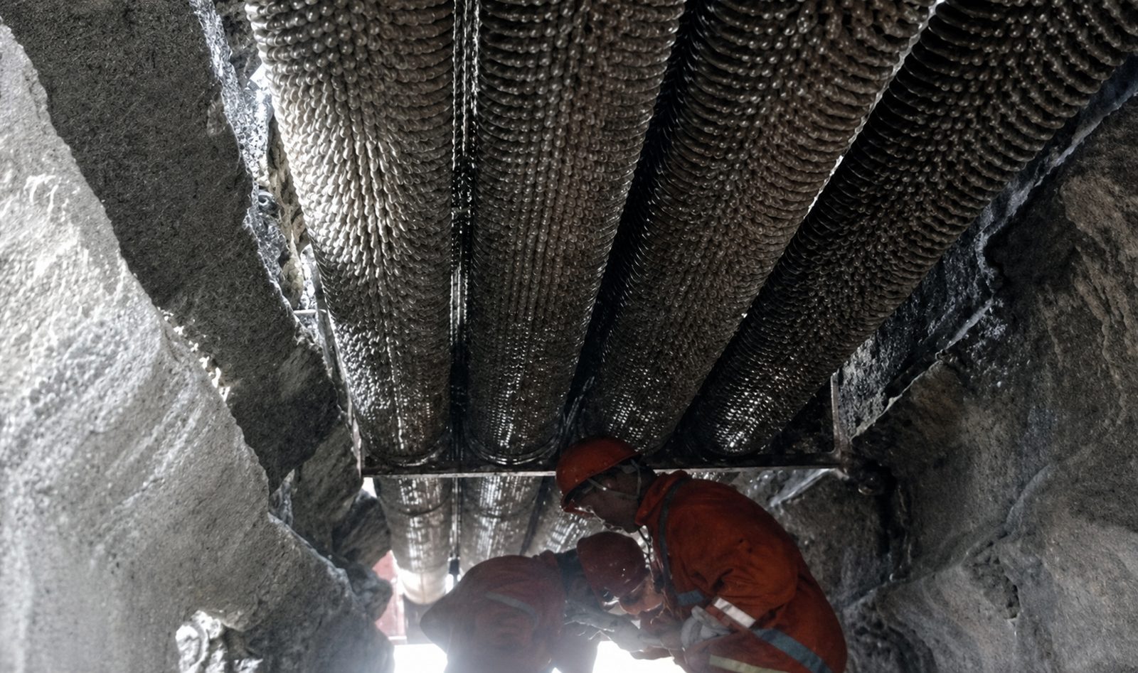 Workers in orange protective gear working inside an industrial heater structure with finned tubes.