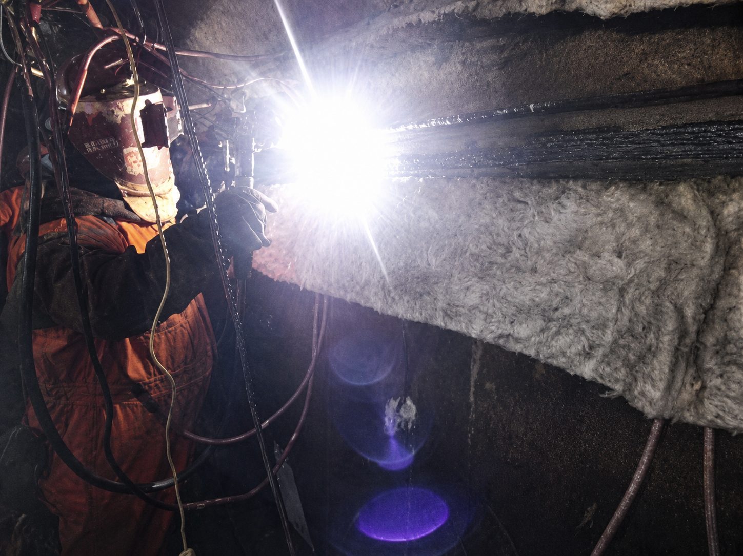 Worker in orange safety gear welding on a metal surface under bright light.